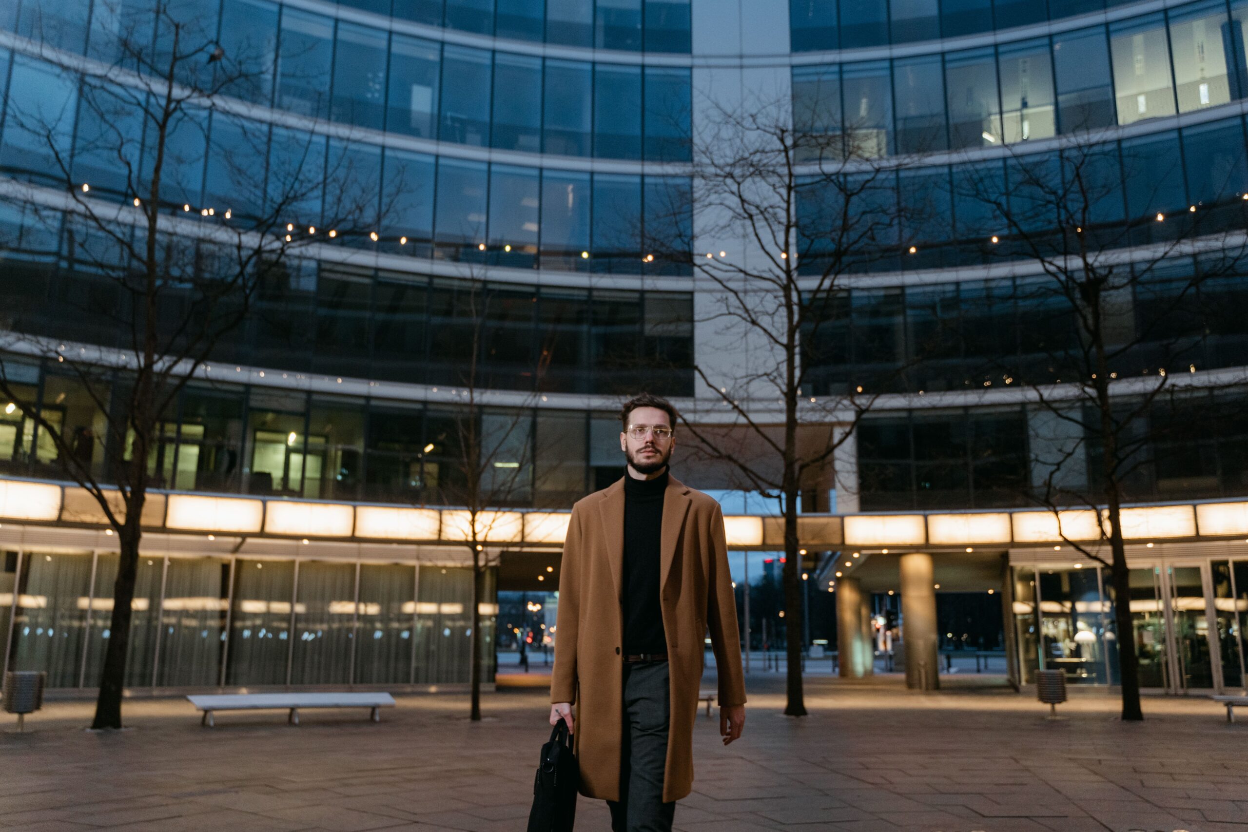 Man in brown coat walking with laptop bag outside a modern glass office building at night.